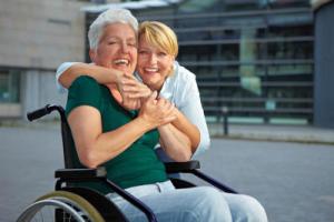 A smiling lady in a wheelchair being hugged by a blonde lady.
