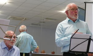 A white-haired bearded man standing behind a music stand with an identical man sitting behind another