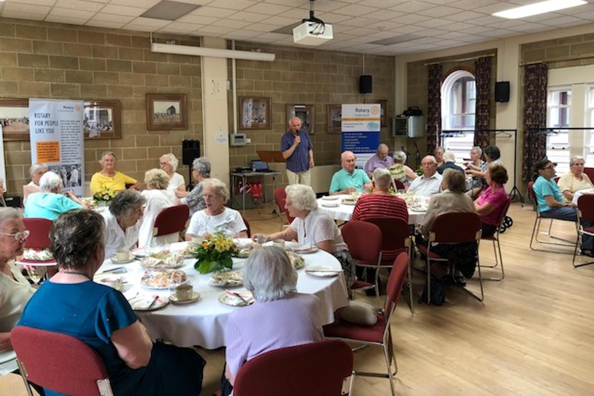 People sitting around dining tables with someone at a microphone.