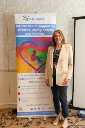 A woman with long blonde hair standing next to a pull-up Kids Inspire banner