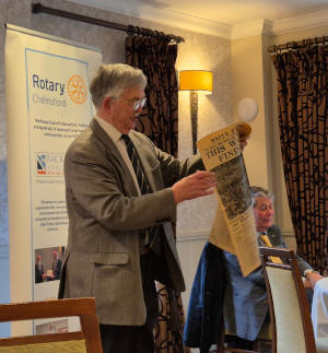 A grey-haired man with glasses and a sports jacket standing in front of a Rotary pull-up banner and looking at a very old newspaper