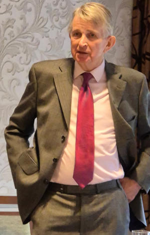 A white-haired man wearing a grey suit, white shirt and red tie