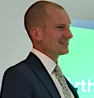 A man with very short hair, wearing a black suit