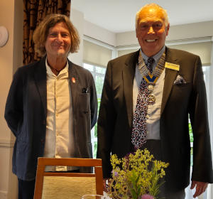 Two men, one wearing a chain of office, standing behind a chair and flower display.