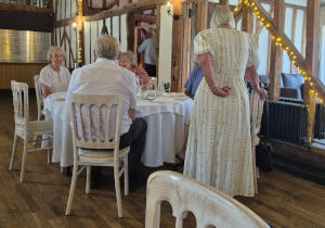 A group of people seated at a circular dining table with a standing woman, with her back to the camera, chats to them