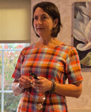 A woman with long dark hair wearing a dress in brown check and holding a pair of glasses.