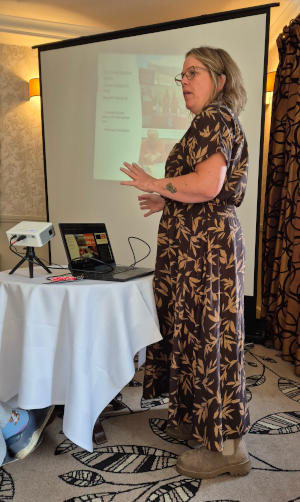A lady with glasses standing to one side of a projector screen, with a laptop and a projector on the table in front of her