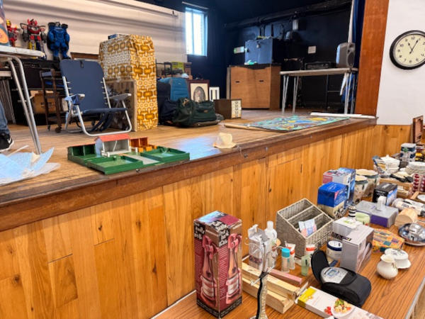 The front of a wooden stage with various things on it and a table in front of it, also with various items