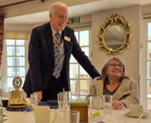 A man standing at the head of a dining table with a lady sitting on his left