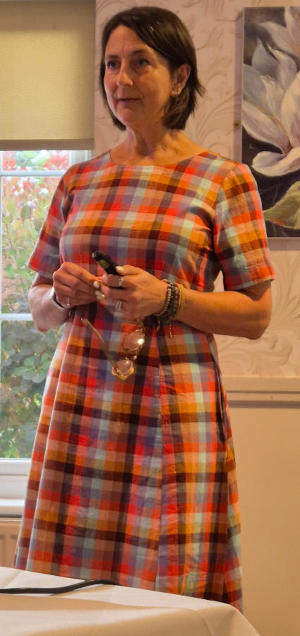 A dark-haired lasdy wearing a brown check dress and holding glasses