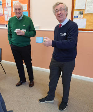 Two tall grey-haired men holding cups standing in front of two notice boards