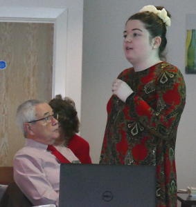 A young woman talking into a microphone with a grey-haired man looking on