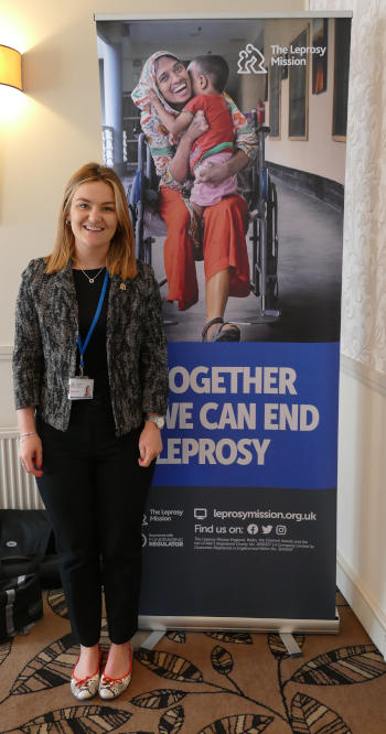 A young woman with long blonde hair standing next to a Leprosy Mission pull-up banner