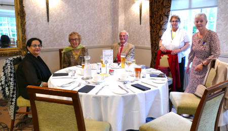 Two women and a man sitting at a dining table with two other women standing next to it