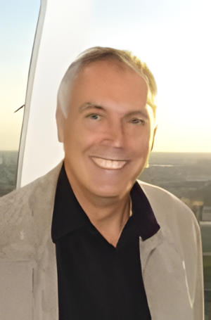 A smiling man wearing a linen jacket over an onpe-necked black shirt