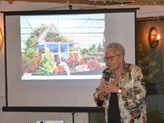 A grey-haired lady holding a microphone and standing in front of a screen showing a picture of a floral display