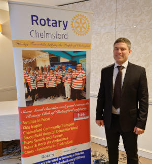 A man wearing a suit standing to the right of a Rotary Club of Chelmsford pull-up banner