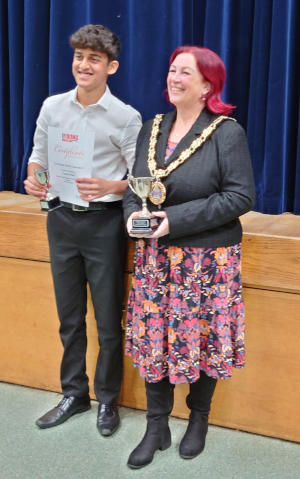 A teenaged male holding a certificate standing next to an older woman wearing a Mayor's chain of office and holding a trophy