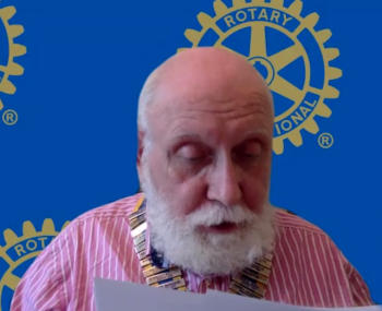 Man with white beard in front of blue background bearing Rotary logos