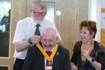 A man in short sleeves putting an office holder's collar around the neck of another man with a woman looking on