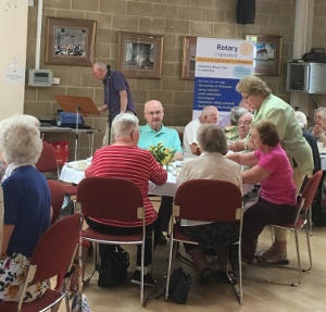 People enjoying a meal with a lady serving and a gentleman looking at a music stand in the background