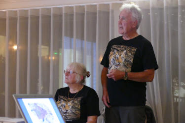 A white-haired couple wearing black T shirts, with a laptop screen nearby