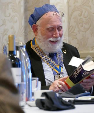 A smiling bearded man in a hat from a cracker with his glasses pushed up to his forehead, wearing a chain of office and looking at a copy of the Shorter Oxford English Dictionary